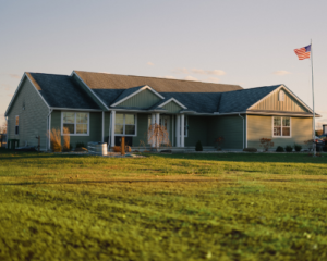 A single-story green house with a gabled roof sits on a vast lawn, under a clear sky. An American flag waves on a tall pole, evoking a serene, patriotic atmosphere.