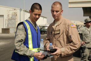 Two men in military attire consult a checklist and walkie-talkie outdoors. One wears a blue safety vest, and another is in a brown uniform. Others stand in the background.